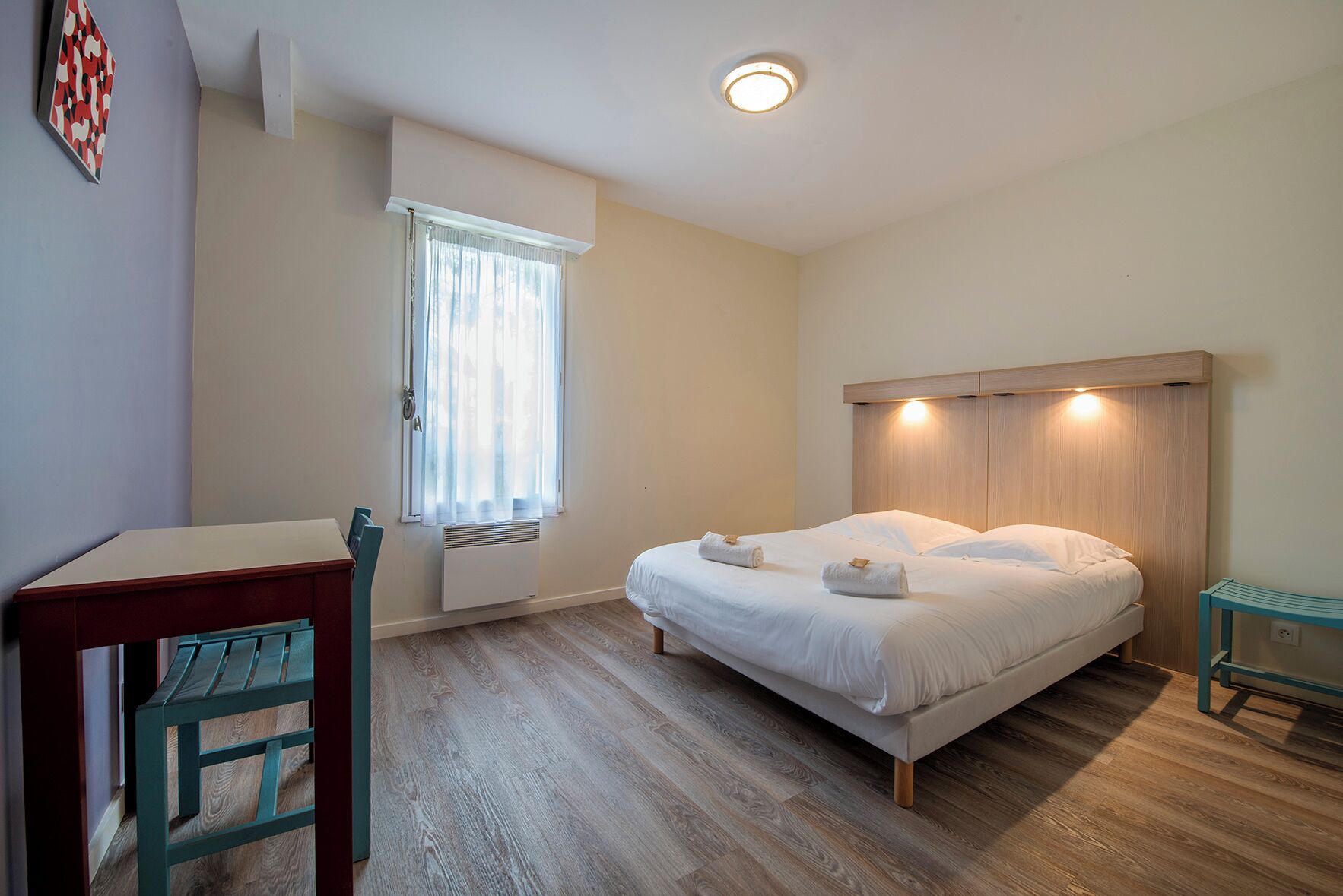 Een slaapkamer met een bed, een bureau, een stoel, een raam met gordijnen en een plafondlamp.