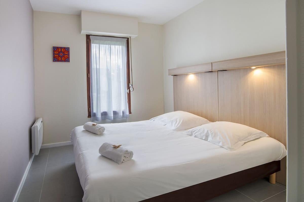 A neatly made bed with two pillows and two rolled towels, a window with sheer curtains, and a wall-mounted radiator.