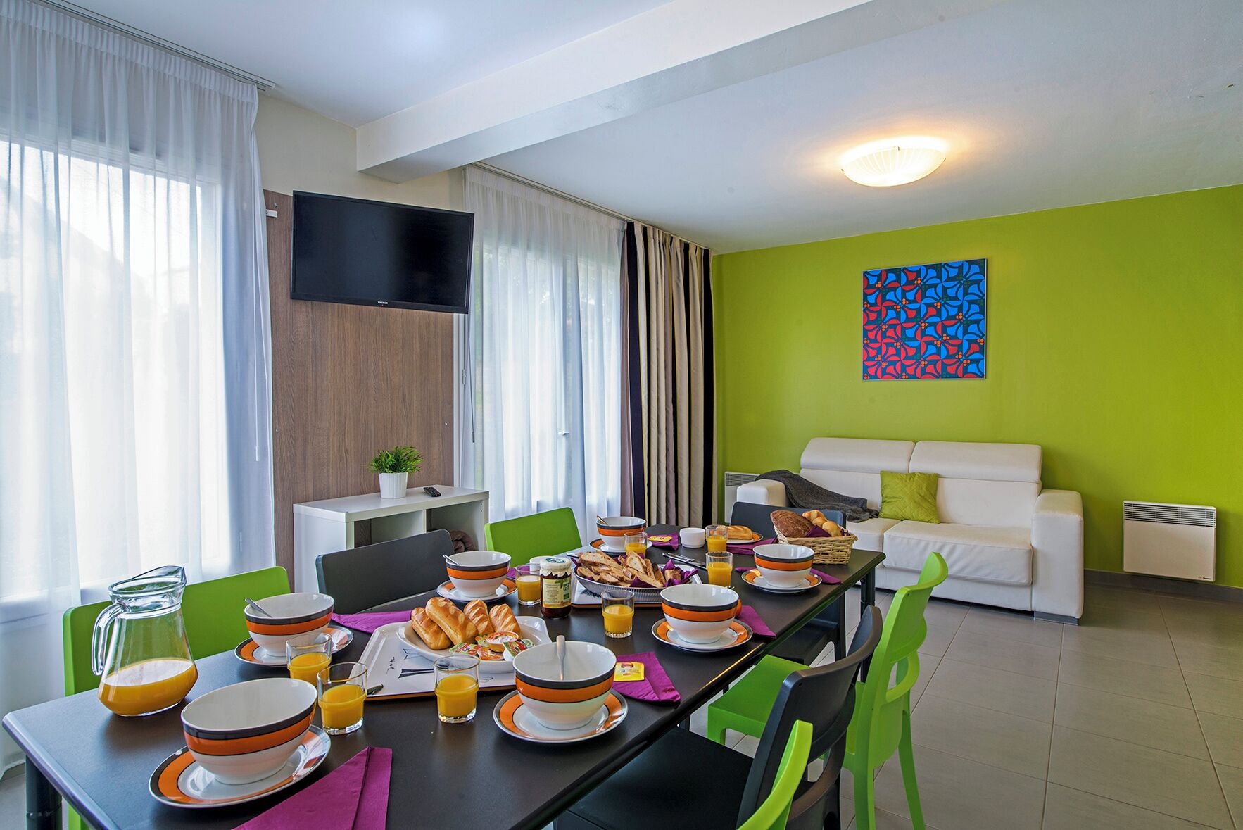 Un comedor con una mesa dispuesta para el desayuno, un sofá blanco y un televisor de pantalla plana montado en la pared.