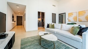 Sala de estar moderna com sofá, mesa de centro e televisão. Uma área de cozinha é visível através de uma porta aberta.