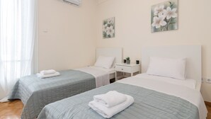 A hotel room with two beds, each with a blue quilted bedspread and white pillows. There is a small bedside table with a lamp, a wall-mounted air conditioner, and two floral paintings on the wall.