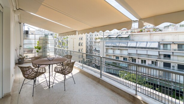 A balcony with a table and chairs, a potted plant, and a view of a cityscape with buildings and a body of water.