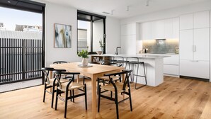 A modern kitchen with white cabinets, a wooden dining table, and black chairs. - Docker St Elwood Apartments by UrbanRest (Elwood)