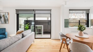 A modern living room with a grey sofa, wooden flooring, and a dining area with a round table and chairs. - Docker St Elwood Apartments by UrbanRest (Elwood)