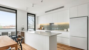 A modern kitchen with white cabinetry, a large island, and a view of the city. - Docker St Elwood Apartments by UrbanRest (Elwood)