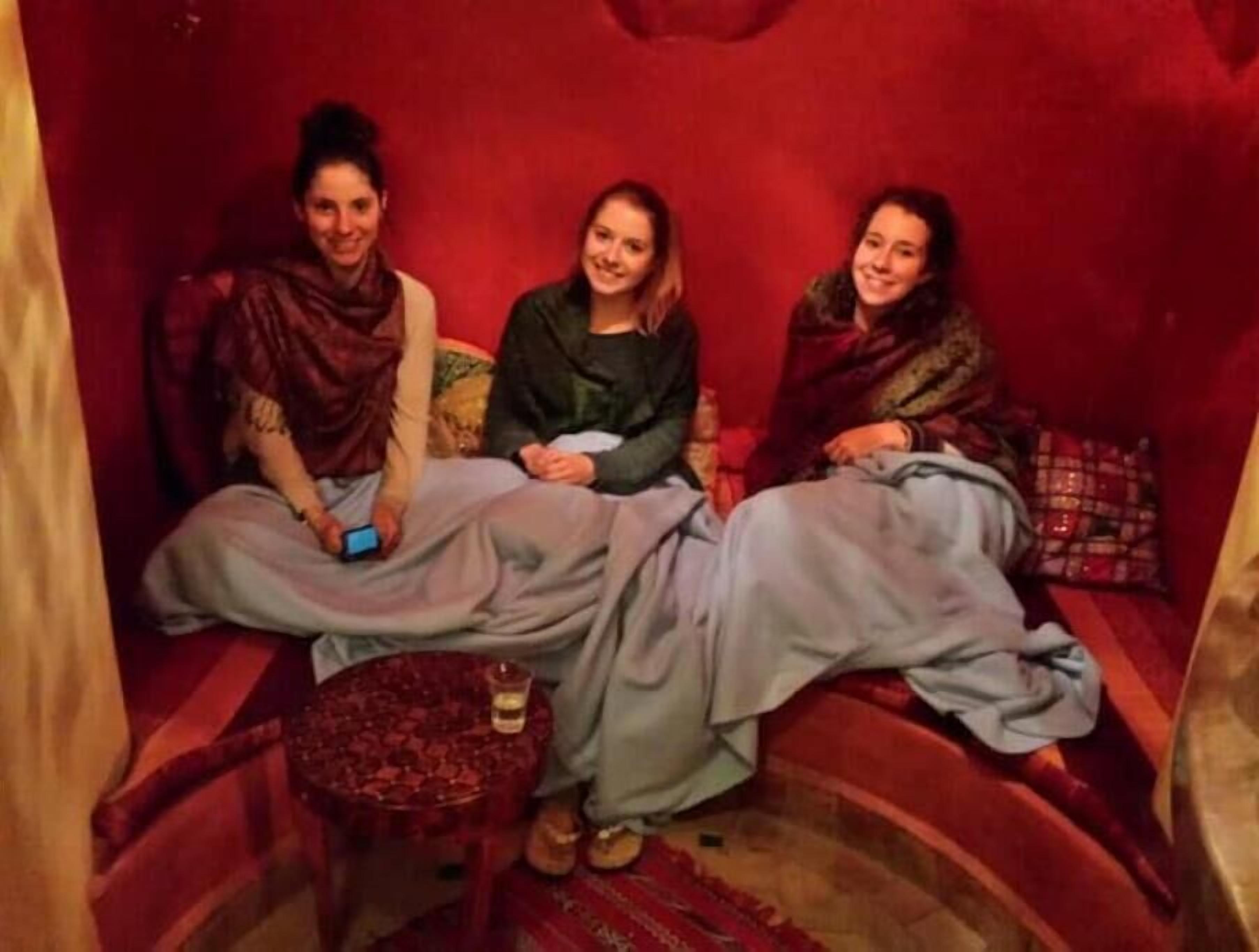 Drie personen zitten op een rode bank in een kamer met een klein rond tafeltje en een fles.