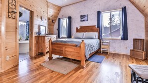 Un dormitorio de cabaña de madera con techo inclinado, una cama de madera, un escritorio y una ventana con cortinas.