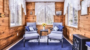 Interior acogedor de una cabaña con paredes de madera, dos sillones grises con cojines azules, una mesita con un jarrón de girasoles y una estufa negra.