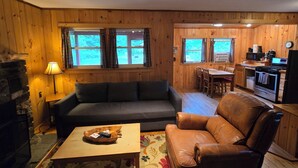 Ein gemütliches Blockhauss-Interieur mit Sofa, Ledersessel, Esstisch, Küchenbereich und Kamin.