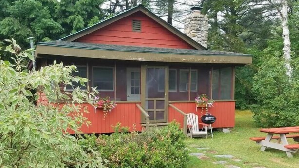 Eine kleine, rote Holzhütte mit Veranda, umgeben von viel Grün und einem Steinsschornstein.