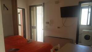 A room with a bed, a table, a washing machine, and a TV. - Trilocale Mimose in Baia Verde (Gallipoli)