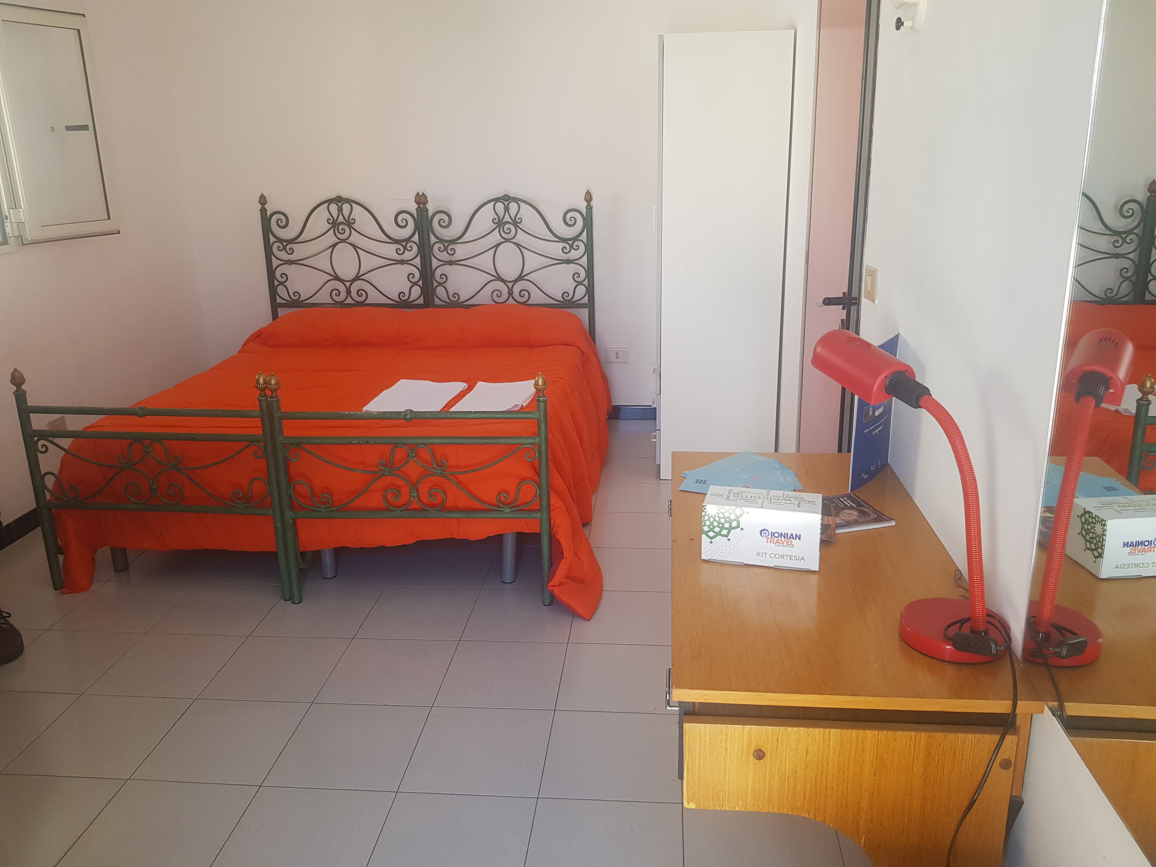 Une chambre avec un lit à ossature métallique, un bureau en bois surmonté d’une lampe rouge et une porte blanche.