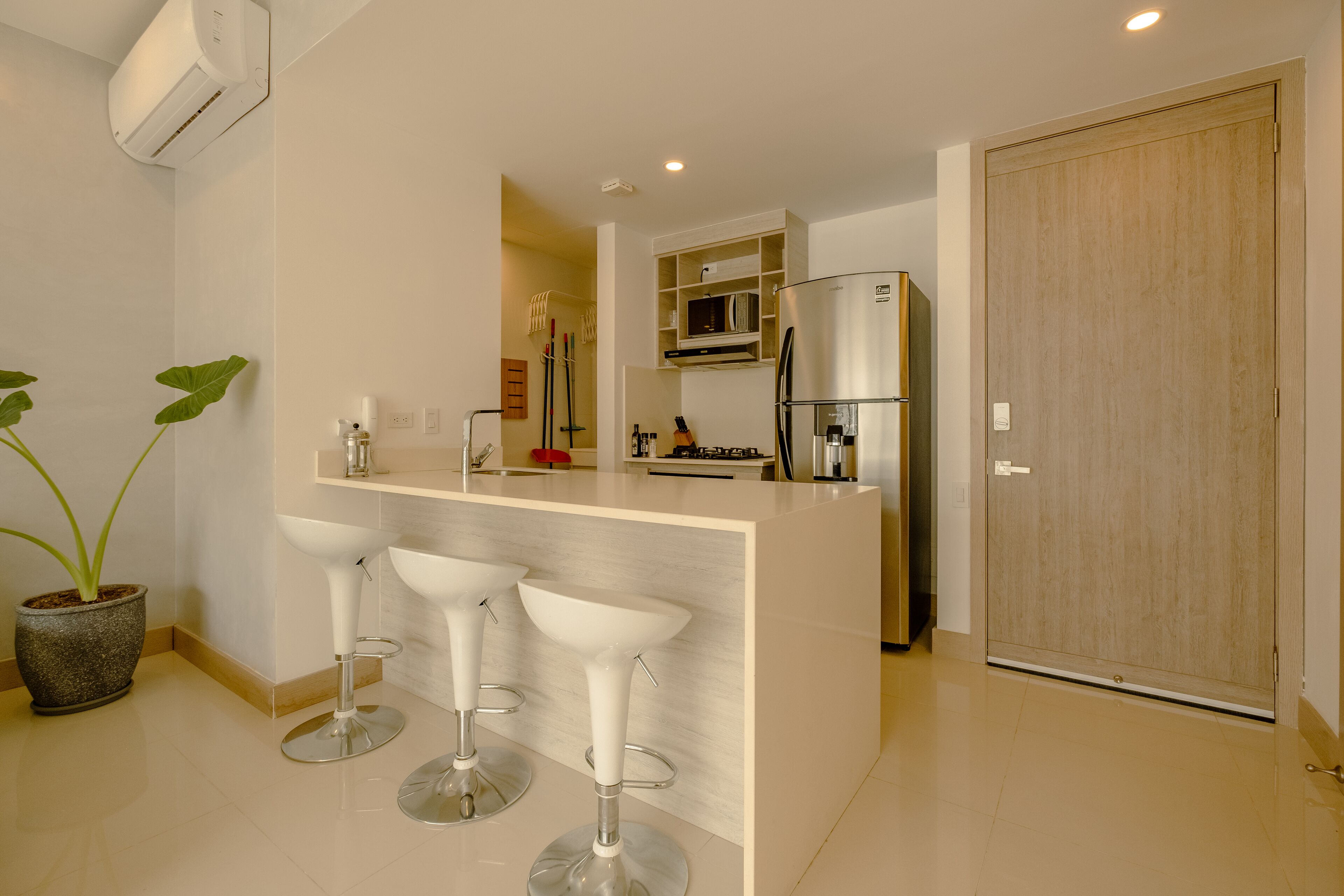A modern kitchen with a central island, bar stools, a potted plant, and a wooden door.