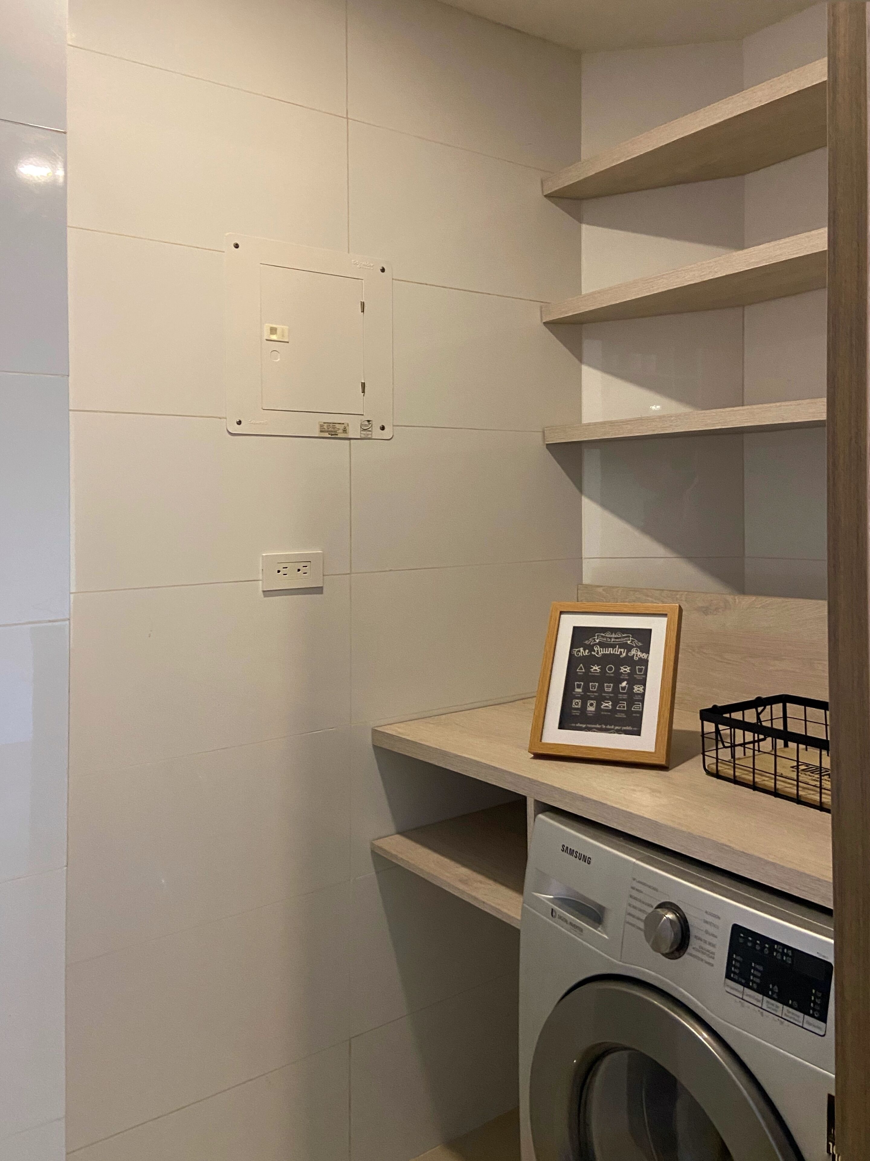 Een wasruimte met een Samsung wasmachine, een lijst met iconen in een kader en een stopcontact op de muur.