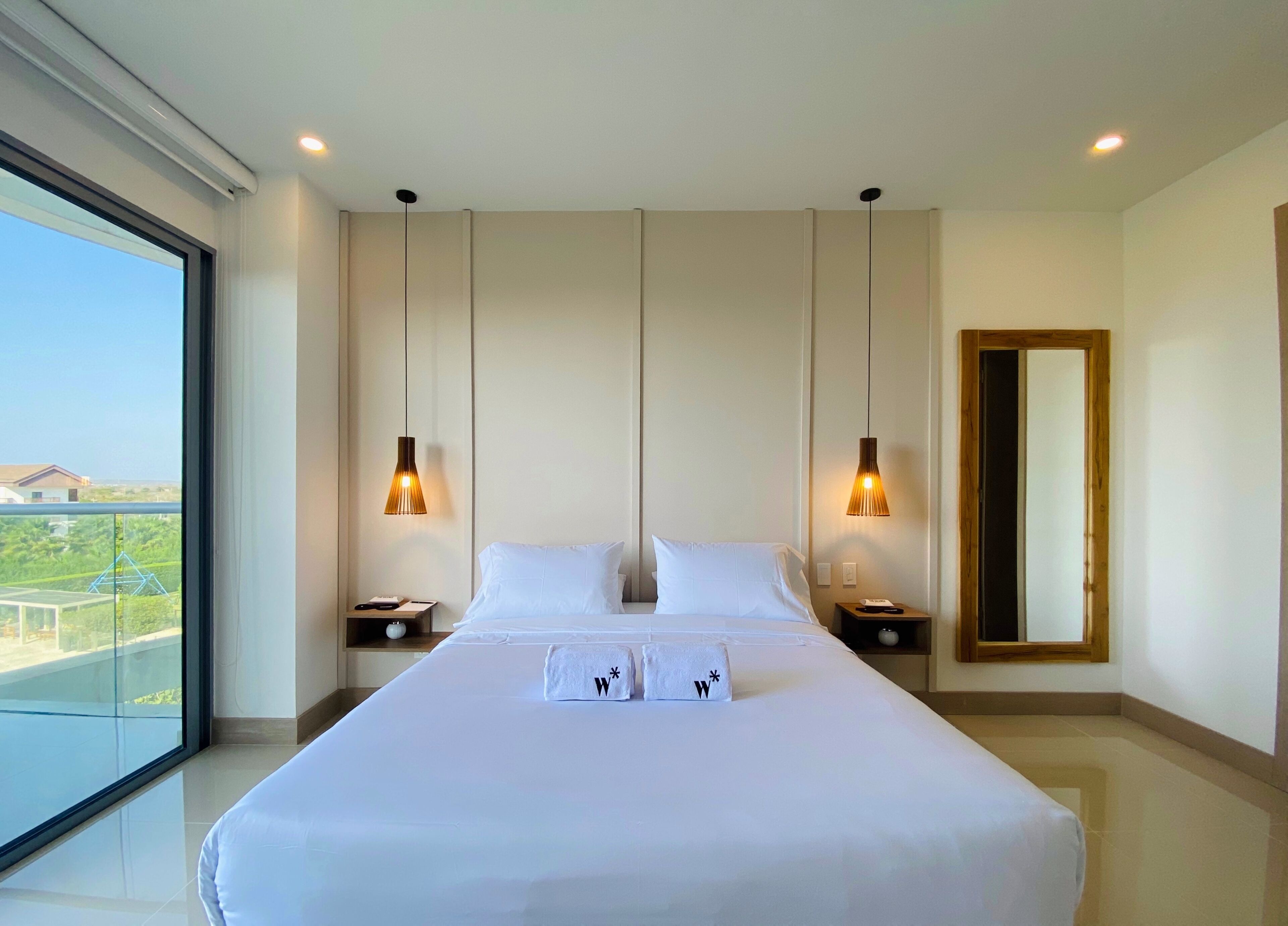 A modern hotel room with a large bed, two bedside tables, a mirror, and a view of greenery outside.