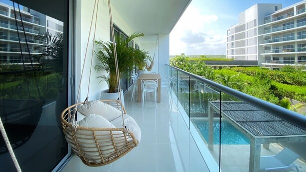 A balcony with a hanging chair, a table with chairs, and a view of a swimming pool and greenery. - Eco by Wynwood House (Cartagena)
