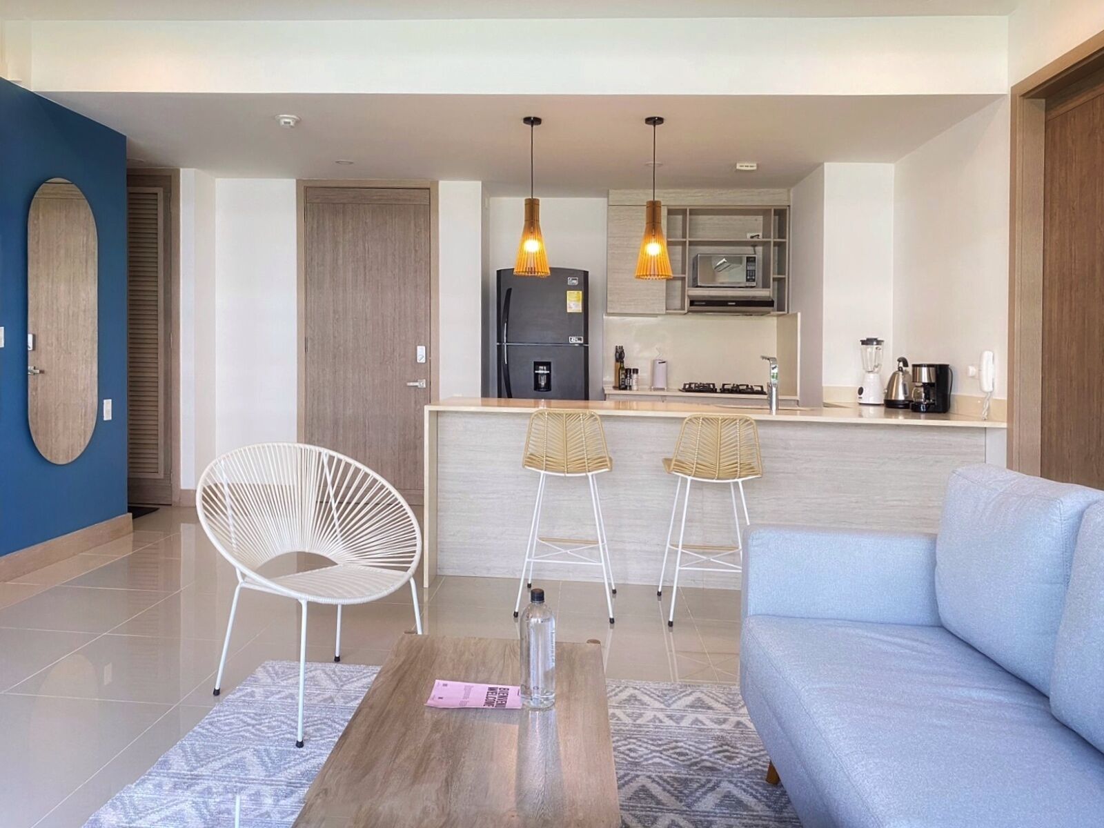 A modern living room with a sofa, a coffee table, and two white chairs. There is a kitchen area with a refrigerator, microwave, and bar stools.
