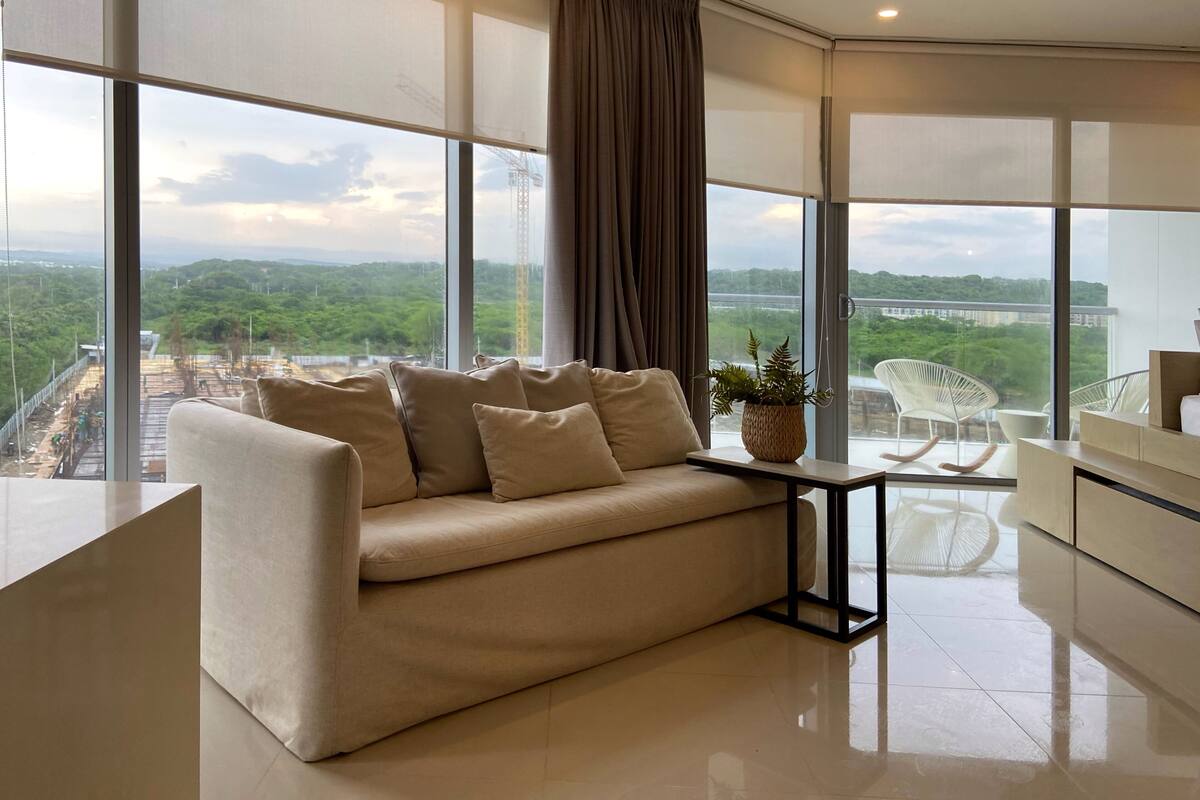 Ein modernes Wohnzimmer mit einem beigen Sofa, einem kleinen Tisch mit Pflanze und Blick auf eine Baustelle durch große Fenster.