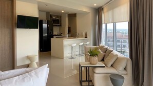A modern living room with a sofa, a coffee table, and a TV. There is a kitchen area with a dining table and chairs. The room has large windows with curtains. - Eco by Wynwood House (Cartagena)