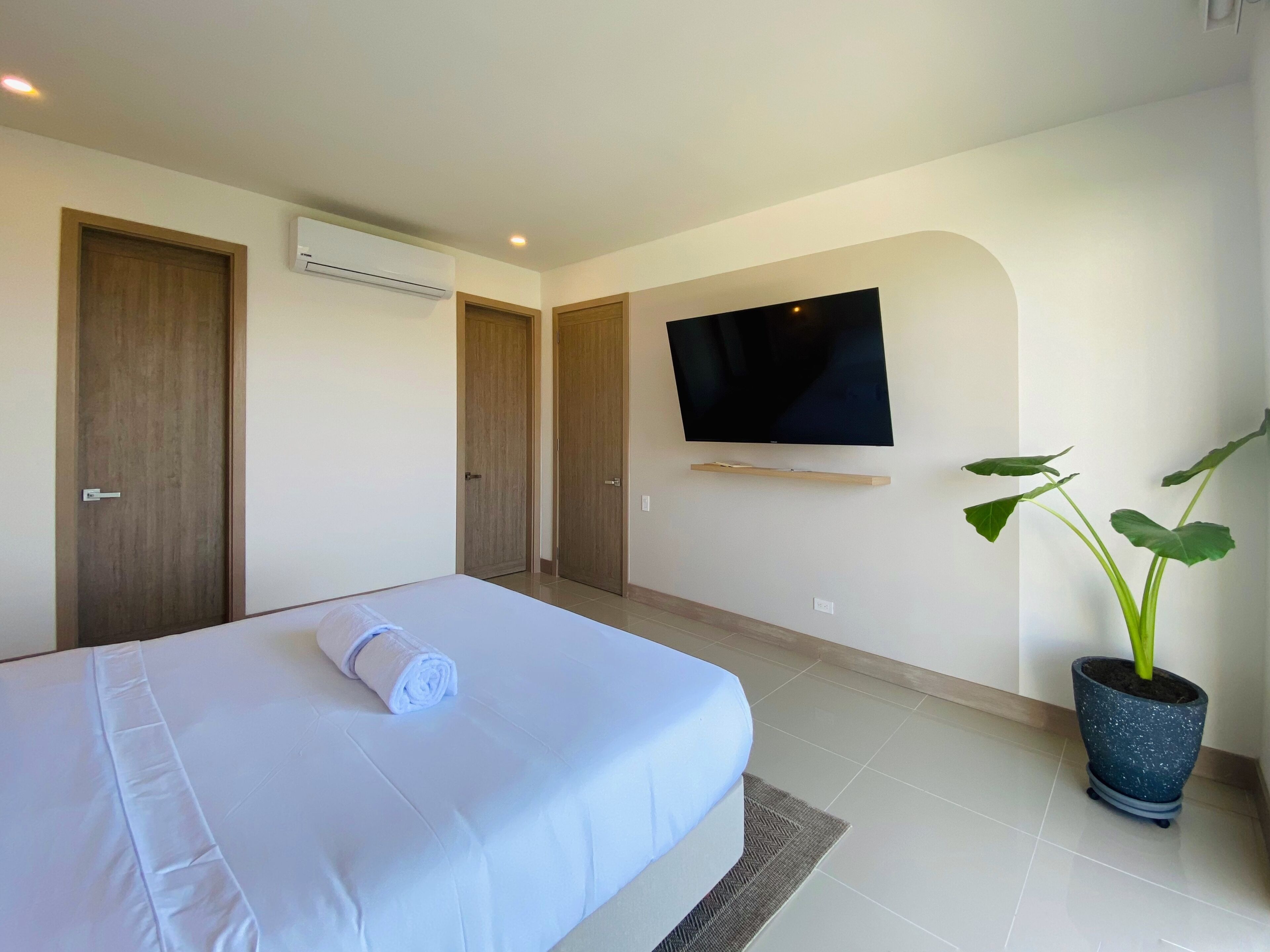 A hotel room with a bed, a flat-screen TV mounted on the wall, a potted plant, and a wooden door.