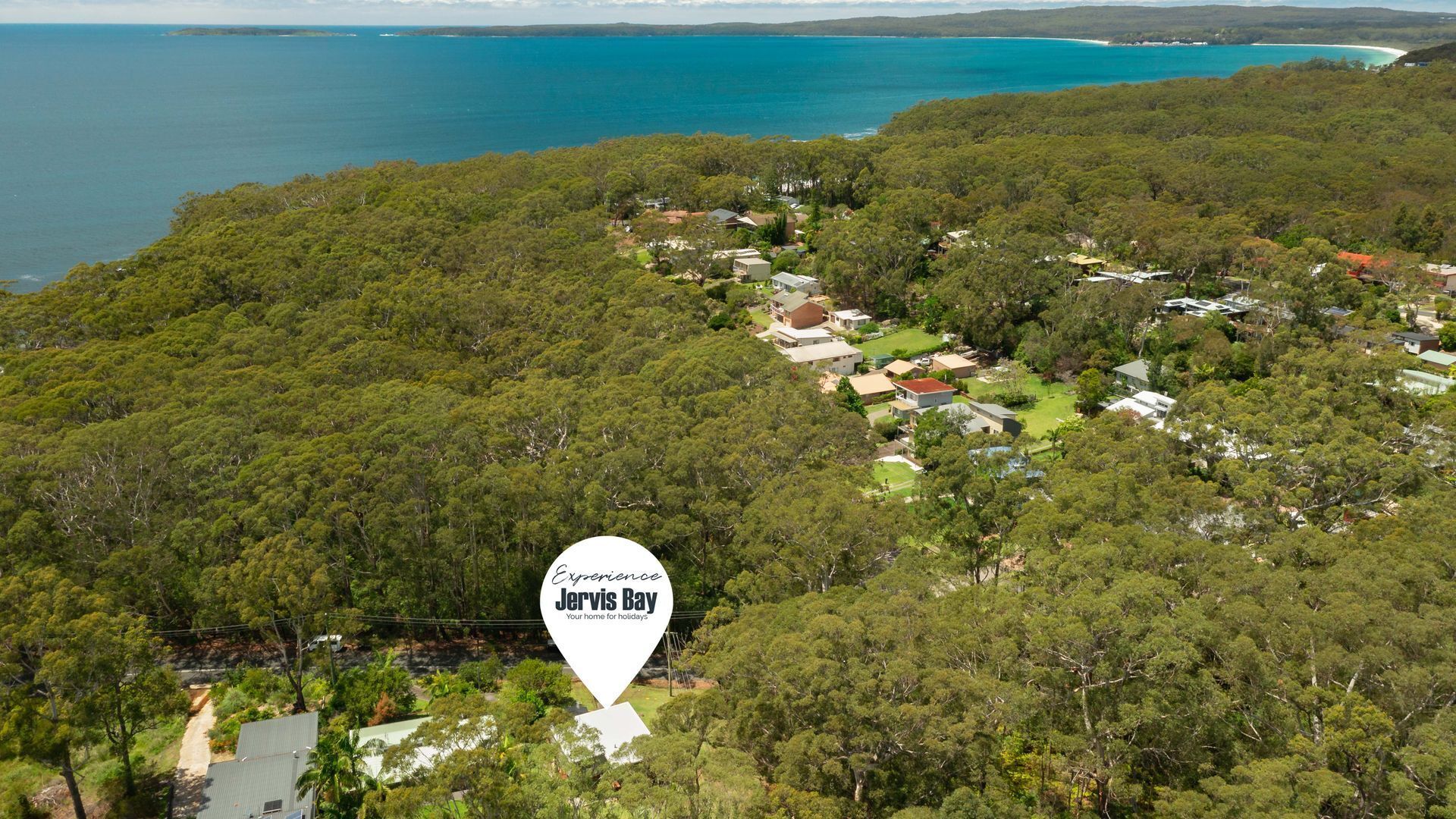 Ett flygbildsfoto av ett bostadsområde med hus omgivna av en tät, grön skog, ett klart blått hav och en vit skylt som visar texten: ”Upplev Jervis Bay – ditt semesterparadis”.