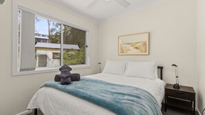 Un dormitorio con un ventanal grande, una cama con ropa de cama blanca, un edredón azul y un cuadro en la pared.