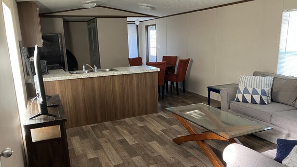 A modern kitchen with a central island, a dining area with red chairs, and a living room with a glass coffee table and a sofa.