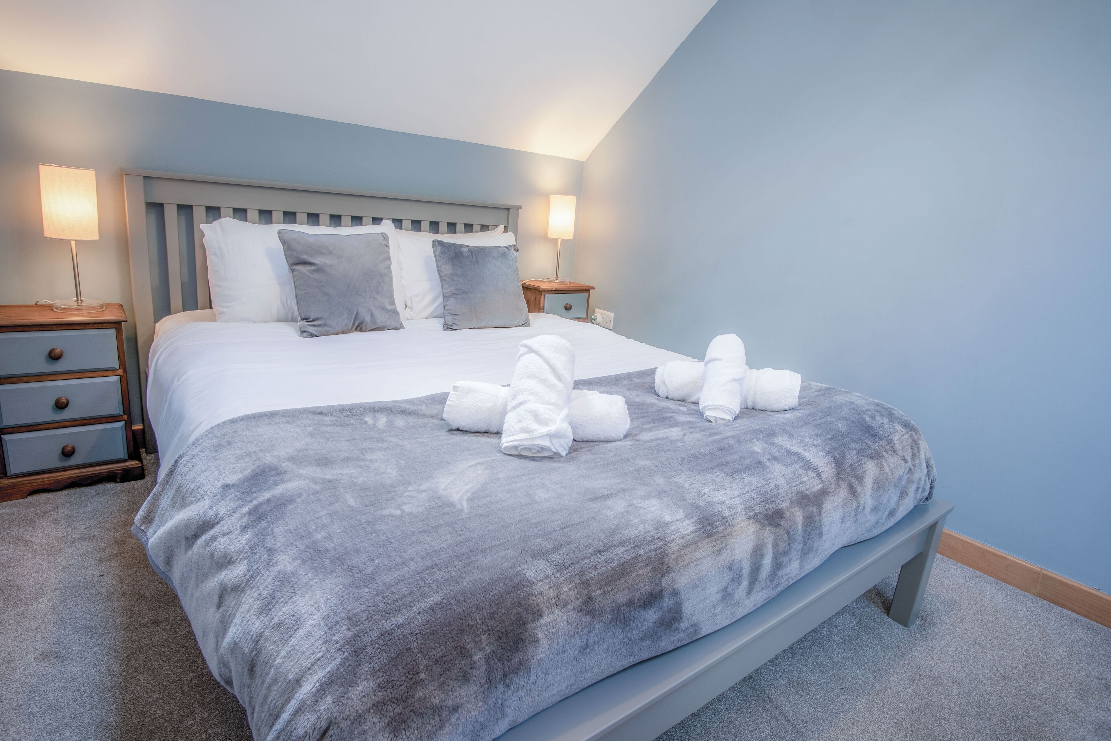 A neatly made bed with a grey blanket and pillows, two bedside tables with lamps, and a wooden bench at the foot of the bed.