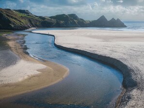 Un paysage côtier avec une plage de sable, un étroit canal d’eau et, au loin, des formations rocheuses.