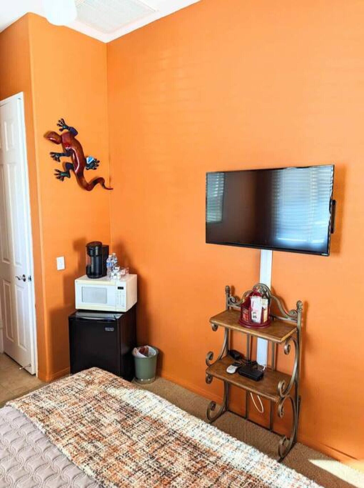 A room with orange walls, a flat-screen TV mounted on the wall, a small wooden table with a microwave and coffee maker, and a bed with a patterned blanket.