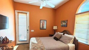 Ein Schlafzimmer mit Bett, Deckenventilator, Tür, Fenster mit Jalousien, Lampe und einem Bild an der Wand.
