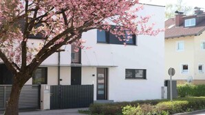 A modern two-story residential building with a gated entrance, a tree with pink blossoms, and a well-maintained lawn. - Apartment Bianca Superior (Salzburg)