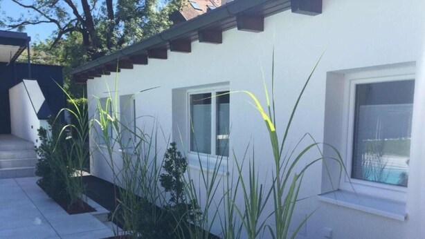 Une maison moderne à l’extérieur blanc, dotée de grandes fenêtres et d’un patio carrelé agrémenté de hautes herbes.