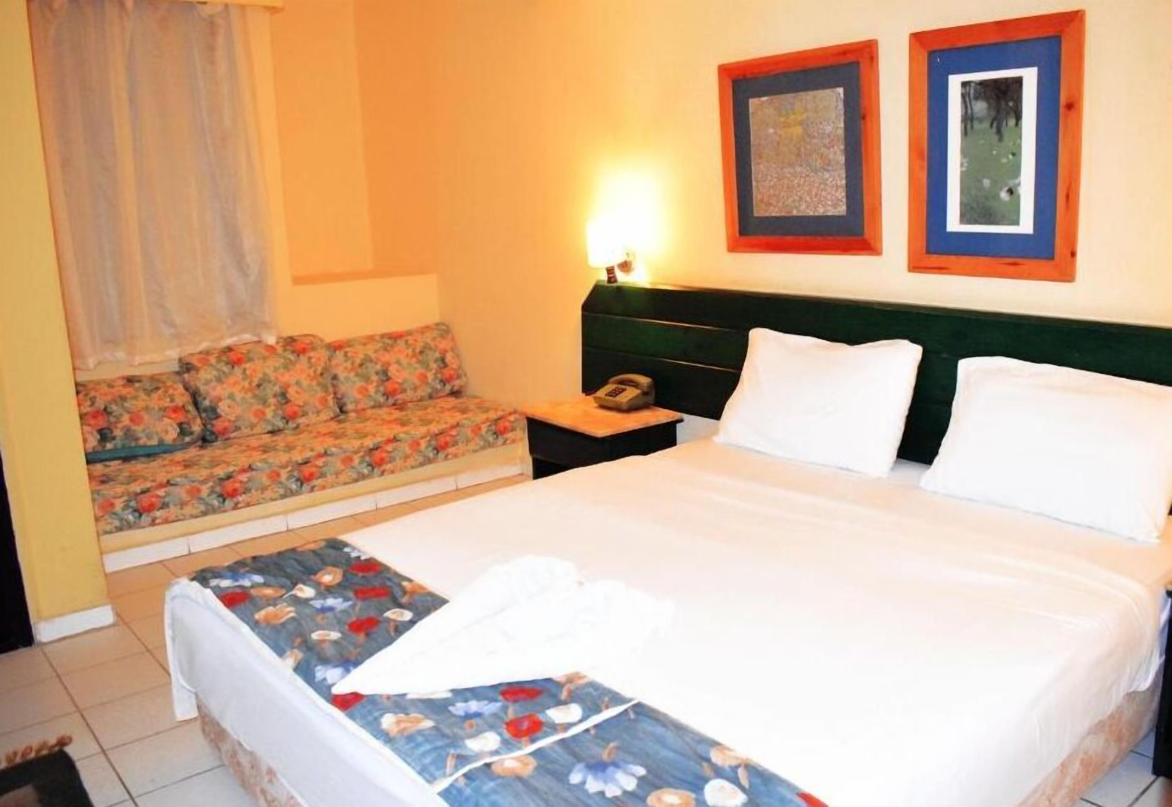 A hotel room with a bed, a sofa, a nightstand with a lamp, and two framed pictures on the wall.