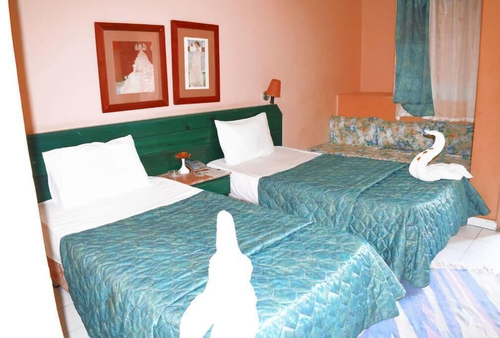 A hotel room with two beds, each with a green bedspread, a nightstand with a lamp, and a framed picture on the wall.