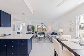 A modern living room with a grey sofa, a coffee table, and a TV. There is a kitchen area with dark blue cabinets and a white countertop.