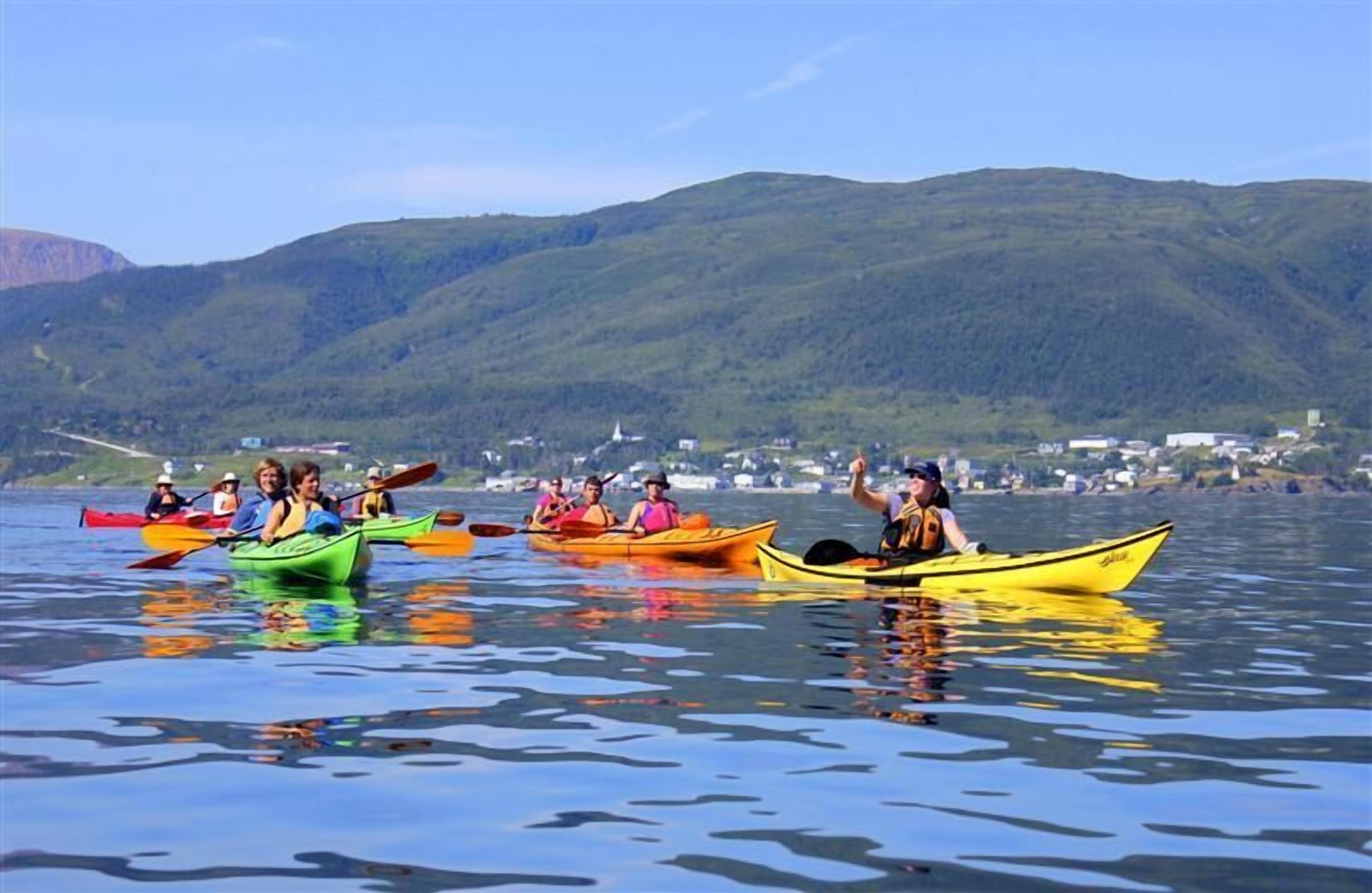 Un groupe de personnes fait du kayak sur un lac paisible, avec une ville en arrière-plan et des montagnes à l’horizon.