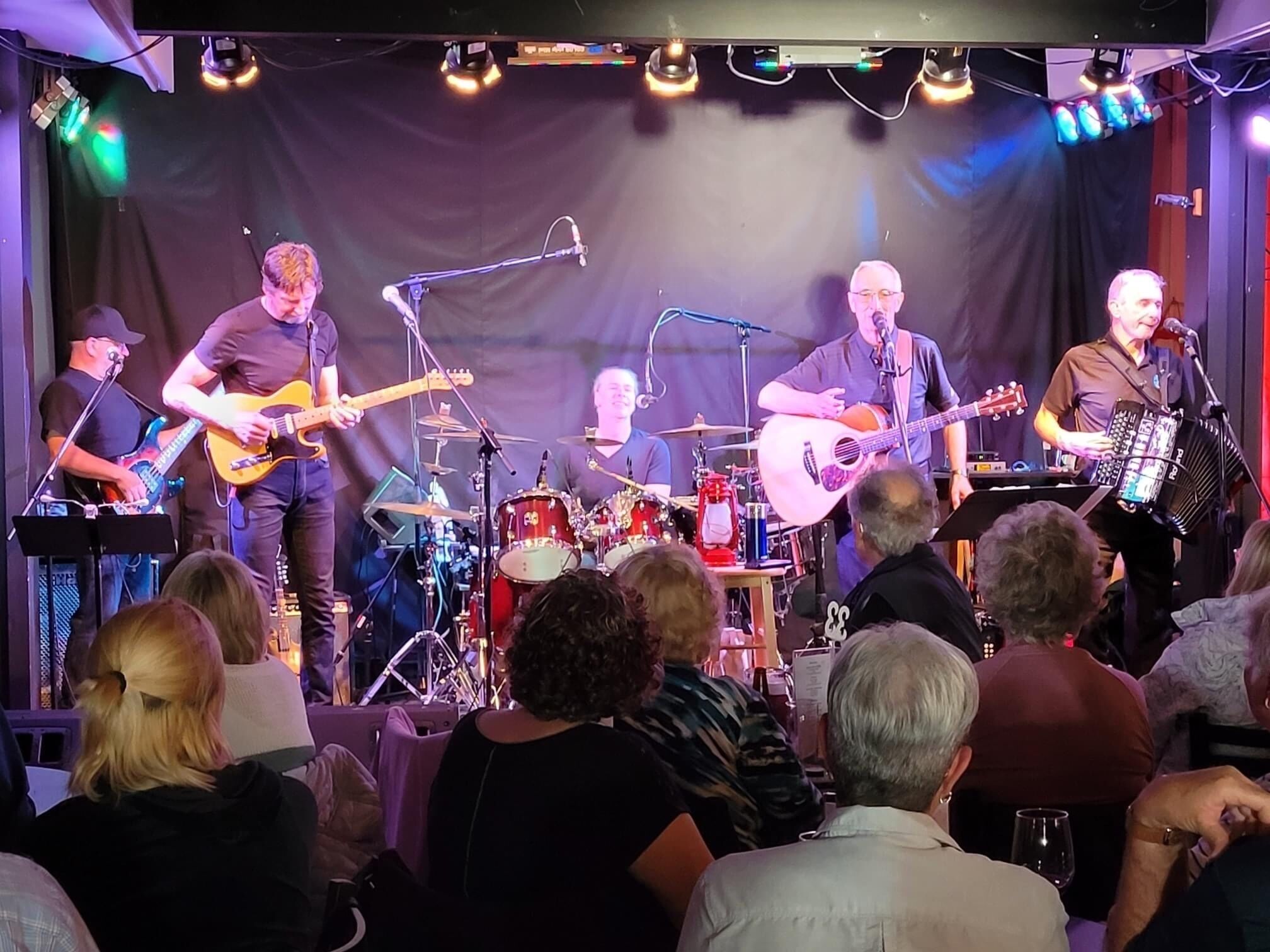 Een band treedt op in de schijnwerpers met een drumstel en diverse instrumenten, een publiek zit vooraan en er staat een microfoonstandaard.
