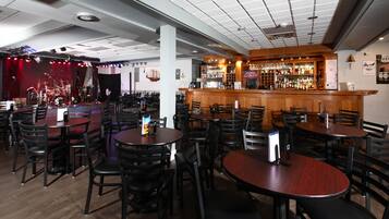 A restaurant with multiple round tables and chairs, a bar area with a wooden counter, and a stage with red curtains in the background.