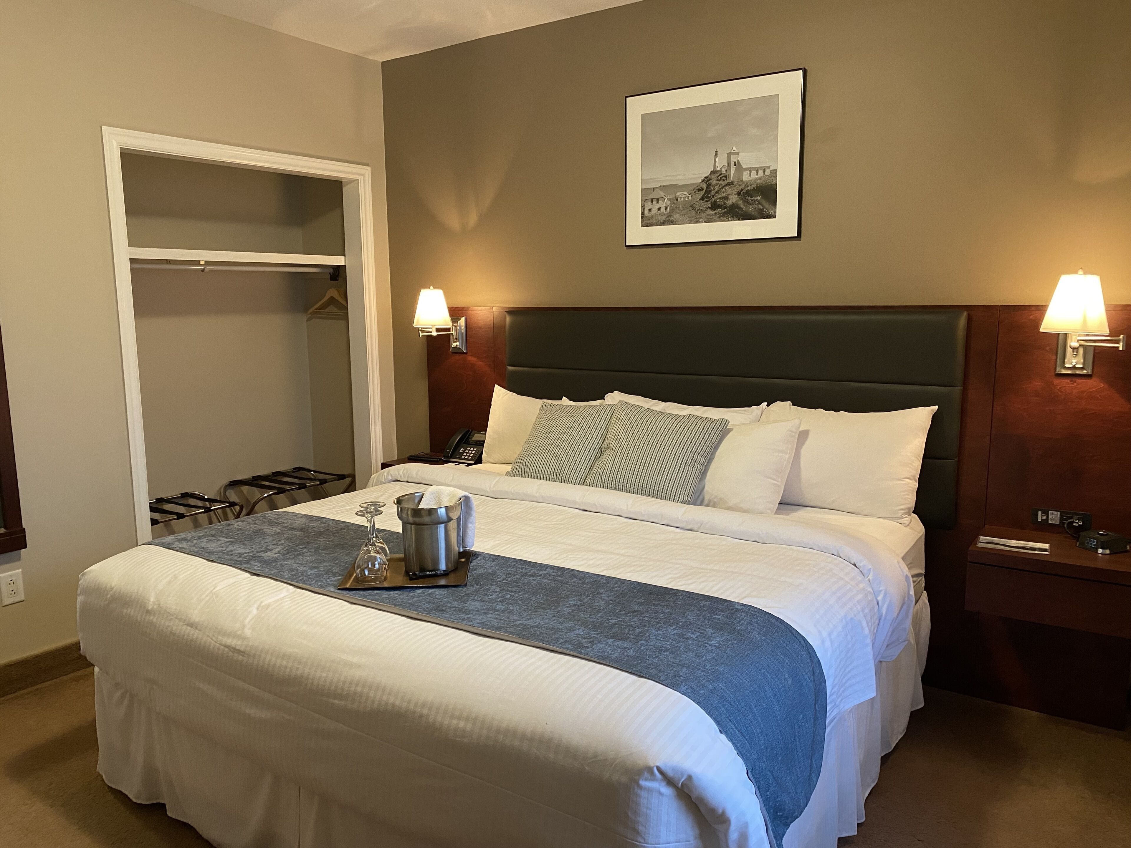 Ein Hotelzimmer mit einem großen Bett, einem Nachttisch, einer Nachttischlampe, einem gerahmten Bild an der Wand und einem Schrank mit Spiegel.