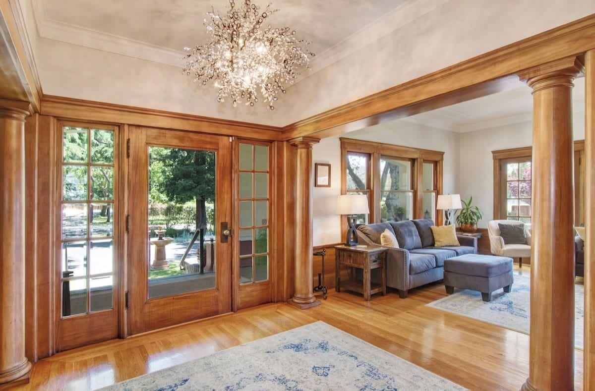 Un salon spacieux agrémenté d’un grand lustre, d’un parquet en bois et offrant une vue sur l’extérieur grâce à de grandes fenêtres.