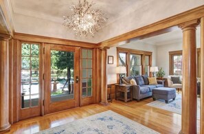 Un salon spacieux agrémenté d’un grand lustre, d’un parquet en bois et offrant une vue sur l’extérieur grâce à de grandes fenêtres.
