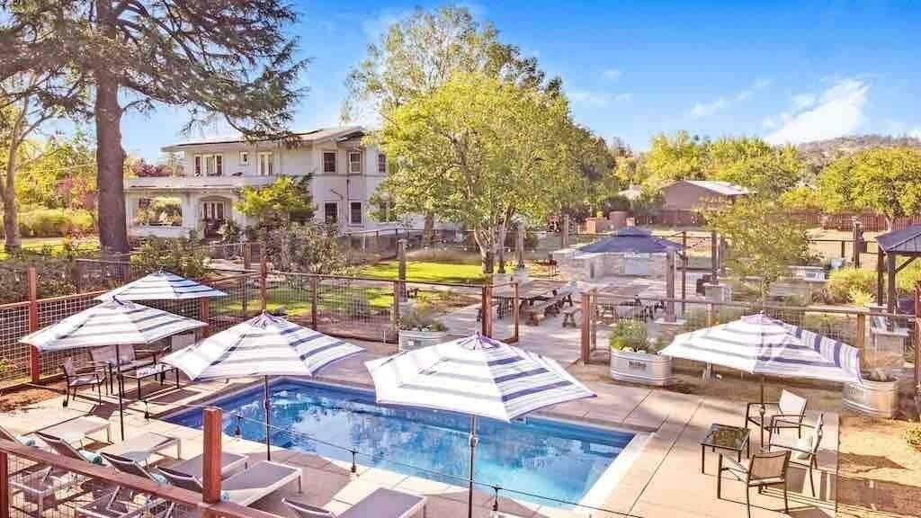 Une piscine entourée de chaises longues et de parasols, elle-même délimitée par une clôture, avec des arbres et un bâtiment visible au loin.