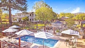 Une piscine entourée de chaises longues et de parasols, elle-même délimitée par une clôture, avec des arbres et un bâtiment visible au loin.