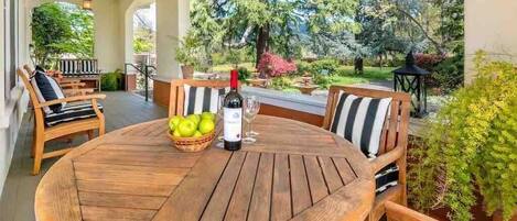 A wooden round table with chairs, a bottle of wine, and a basket of apples on a patio with a view of trees and a garden.