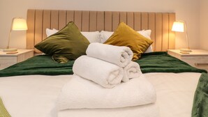 Ein ordentlich bezogenes Bett mit einer grünen Tagesdecke, weißen Kissen und aufgerollten weißen Handtüchern. An beiden Seiten des Bettes stehen Nachttischlampen.