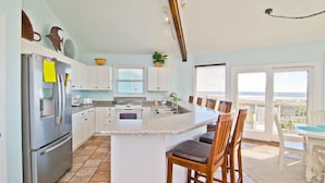 A kitchen with a central island, white cabinets, a stainless steel refrigerator, and a dining area with a view. - Viewpoint, Oceanfront House in Emerald Isle, NC! (Emerald Isle)