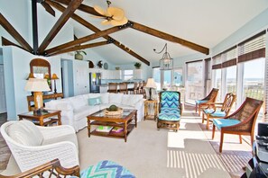 A spacious living room with a high ceiling, wooden beams, a white sofa, a patterned armchair, and a dining area with wooden chairs.
