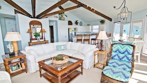 A spacious living area with a white sectional sofa, a wooden coffee table, and a wicker chair. A kitchen with white cabinets and a dining area with white chairs are visible in the background. - Viewpoint, Oceanfront House in Emerald Isle, NC! (Emerald Isle)