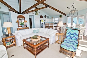 A spacious living area with a white sectional sofa, a wooden coffee table, and a wicker chair. A kitchen with white cabinets and a dining area with white chairs are visible in the background.